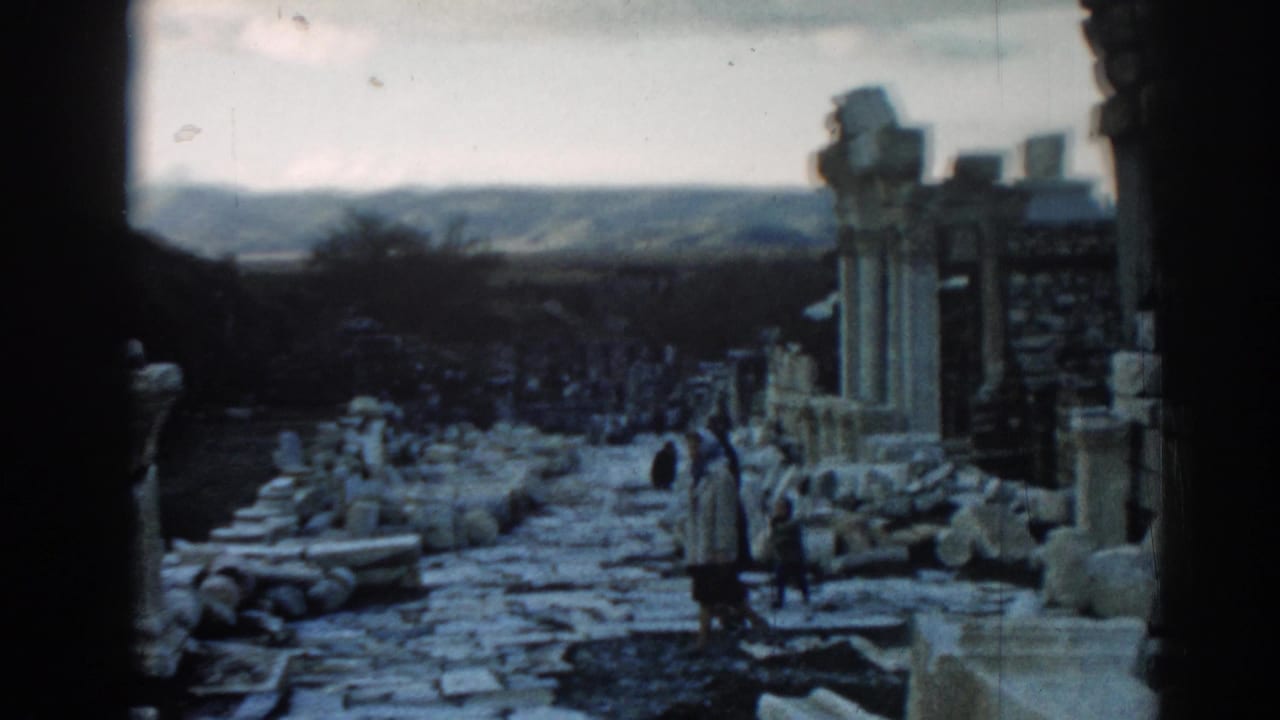 Visitors in coats appear on the Ephesus stone path, surrounded by columns, rubble, and a dark architectural edge.