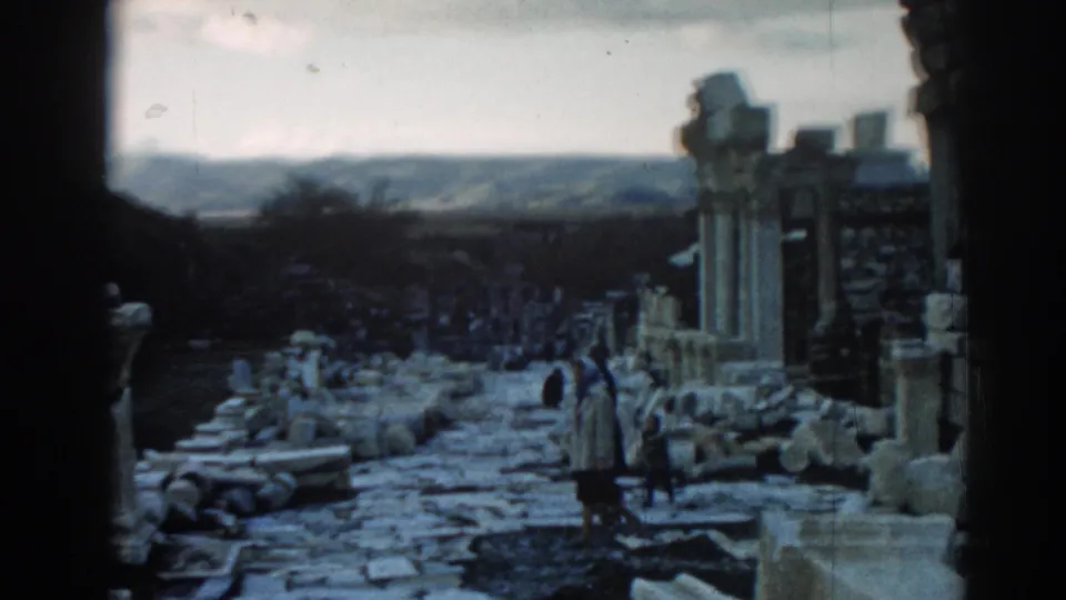 Visitors in coats appear on the Ephesus stone path, surrounded by columns, rubble, and a dark architectural edge.
