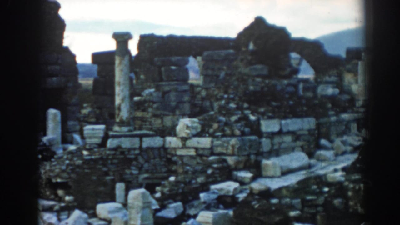 1962 Ephesus travel footage frame with low ruined walls, scattered masonry, upright stone columns, and hills beyond.