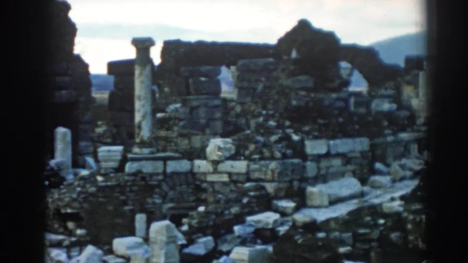 1962 Ephesus travel footage frame with low ruined walls, scattered masonry, upright stone columns, and hills beyond.