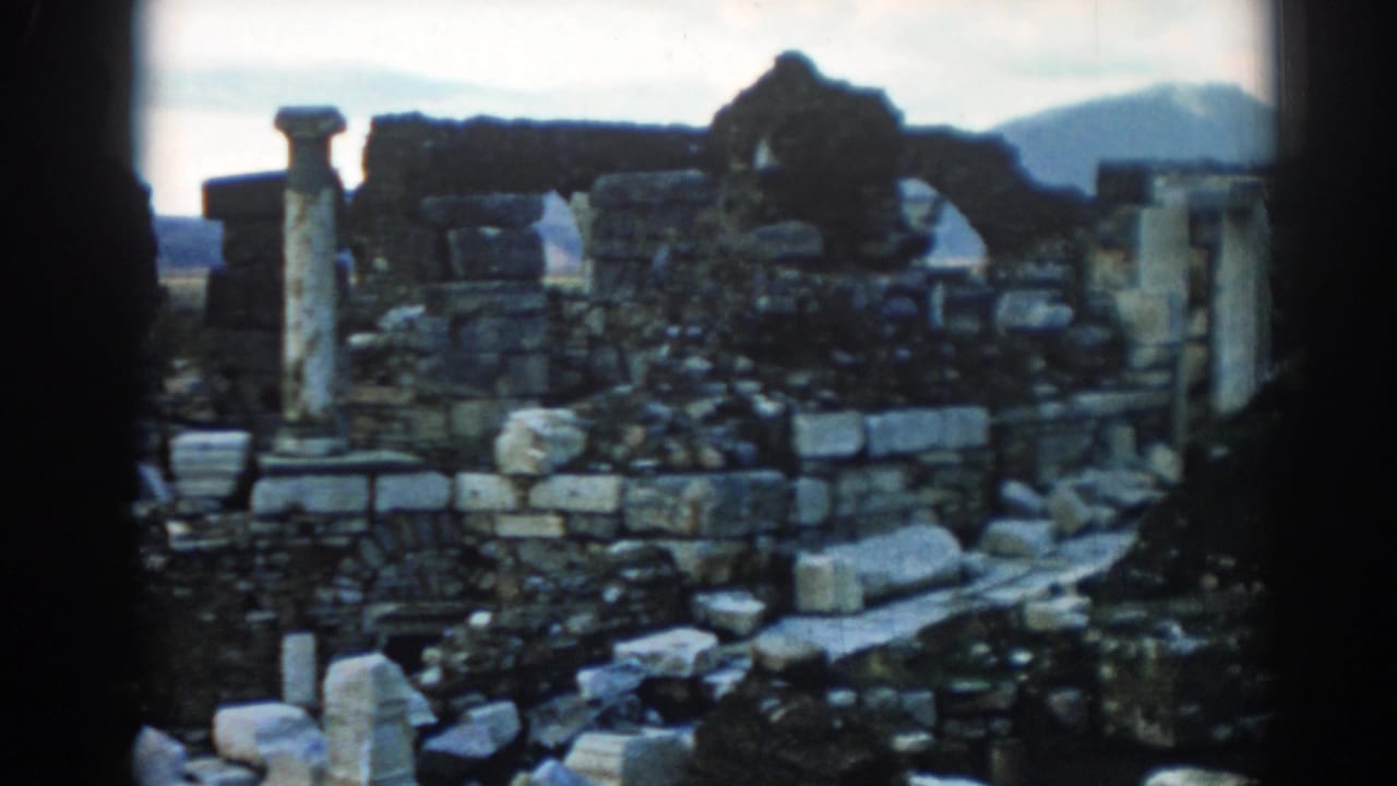 1962 8mm view at Ephesus showing rubble, stacked stone blocks, standing columns, and a hazy hill behind the site.