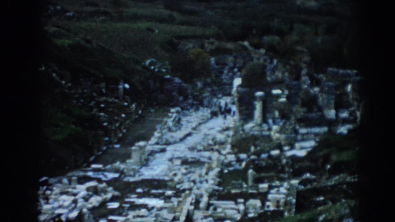 1962 8mm view of Ephesus ruins in Turkey, with stone columns, scattered blocks, and small visitors along a pale pathway.