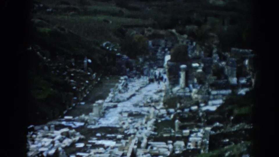 1962 8mm view of Ephesus ruins in Turkey, with stone columns, scattered blocks, and small visitors along a pale pathway.