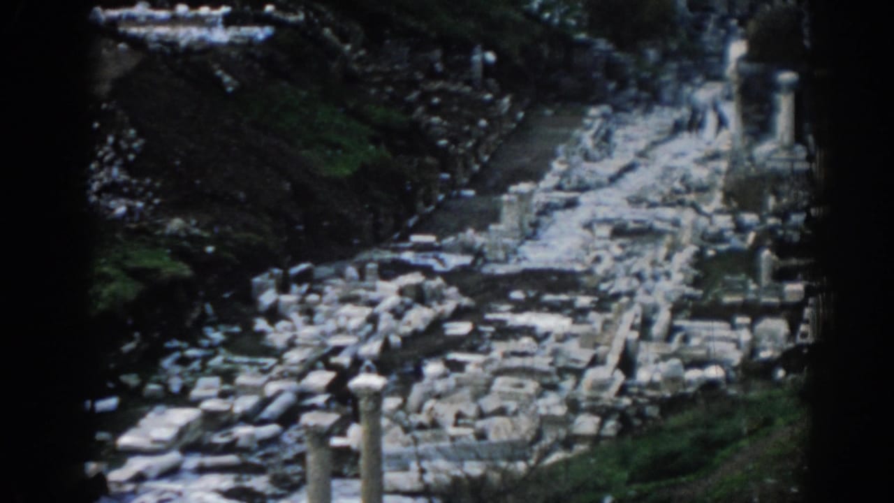 1962 visitor-shot 8mm footage of Ephesus ruins, with a pale route, column remains, scattered stones, and hillside vegetation.