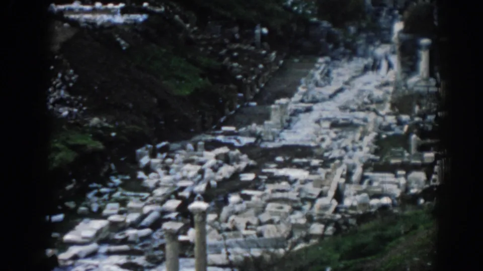1962 visitor-shot 8mm footage of Ephesus ruins, with a pale route, column remains, scattered stones, and hillside vegetation.