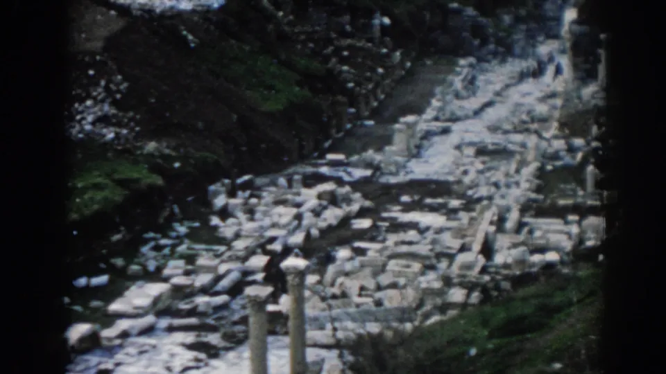 1962 Ephesus travel footage showing a visitor among scattered columns, stone blocks, and grassy embankments.