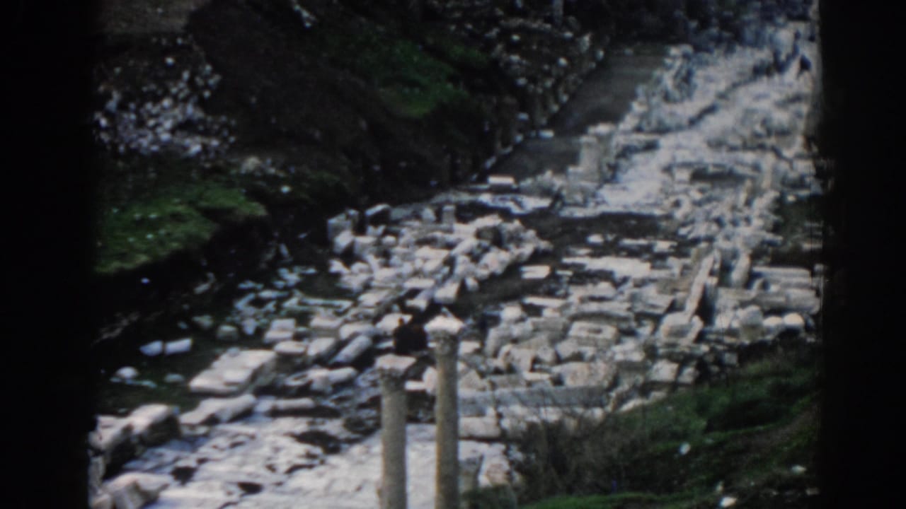 Ancient stone path with scattered blocks, columns, and a distant visitor near the ruins.