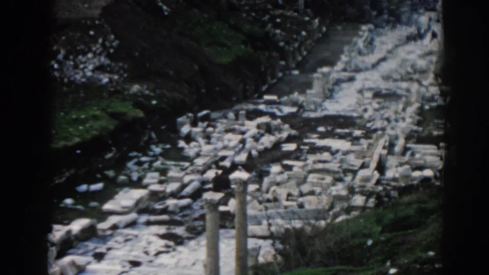 Ephesus ruins in 1962 with a distant visitor, broken stonework, and grassy slopes around the archaeological path.