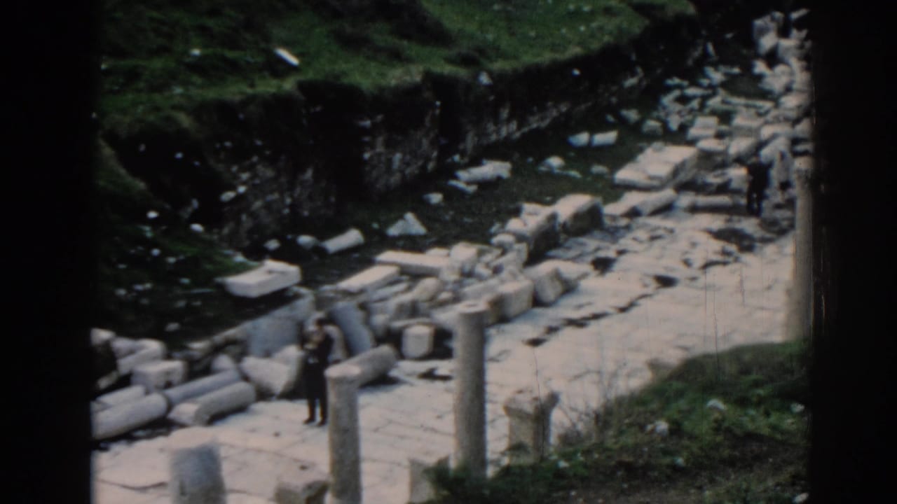 Visitors stand among stone blocks and columns at an ancient ruin site.