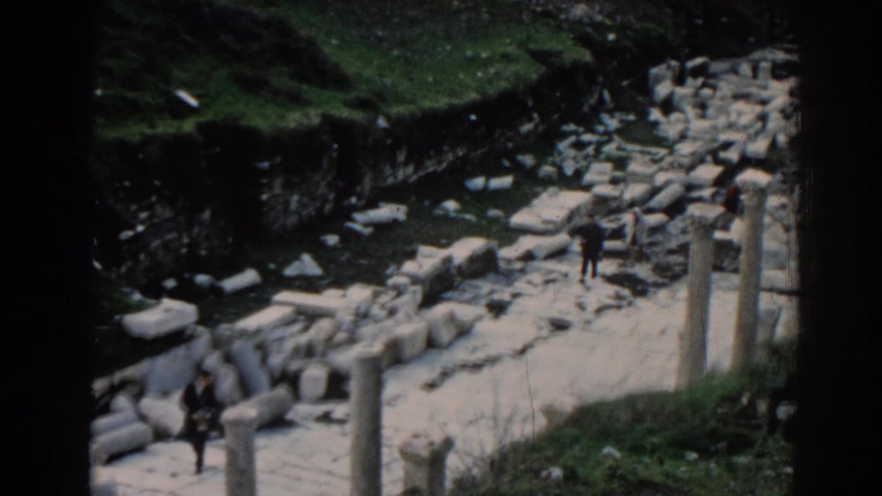 1962 Ephesus footage frame with tourists crossing a ruin path between columns, fallen blocks, and grassy slopes.