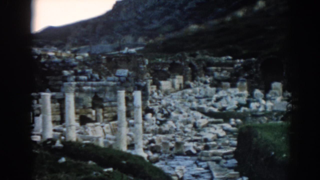 1962 home-movie frame of Ephesus, with a rubble-strewn path, standing columns, and dark gate edges from the film scan.