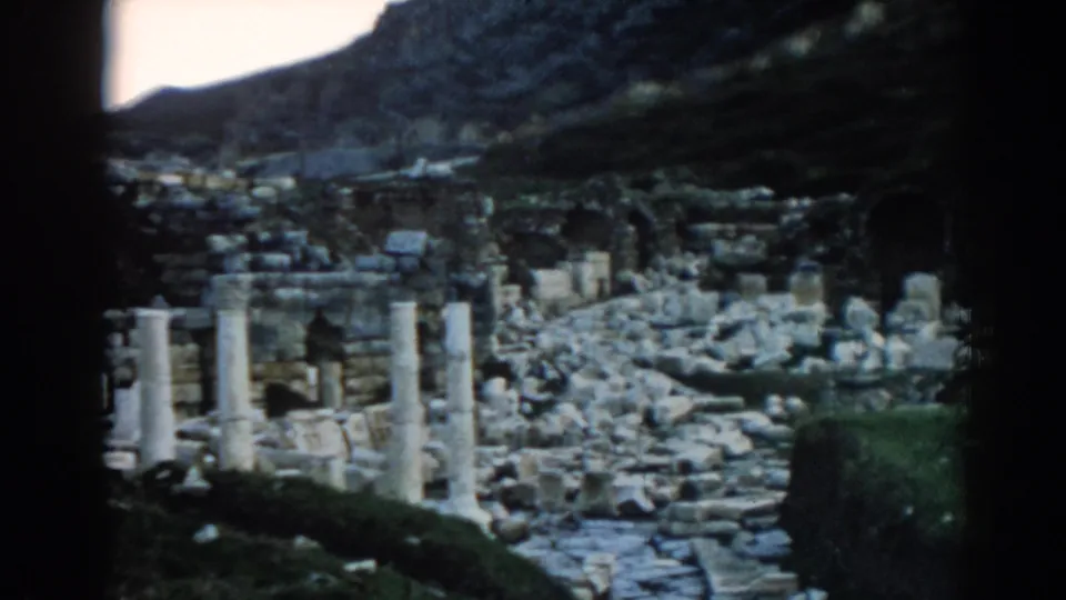 1962 home-movie frame of Ephesus, with a rubble-strewn path, standing columns, and dark gate edges from the film scan.