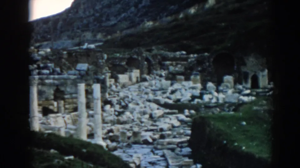 1962 8mm view of Ephesus ruins with scattered masonry, upright columns, arched openings, and a hillside behind the site.