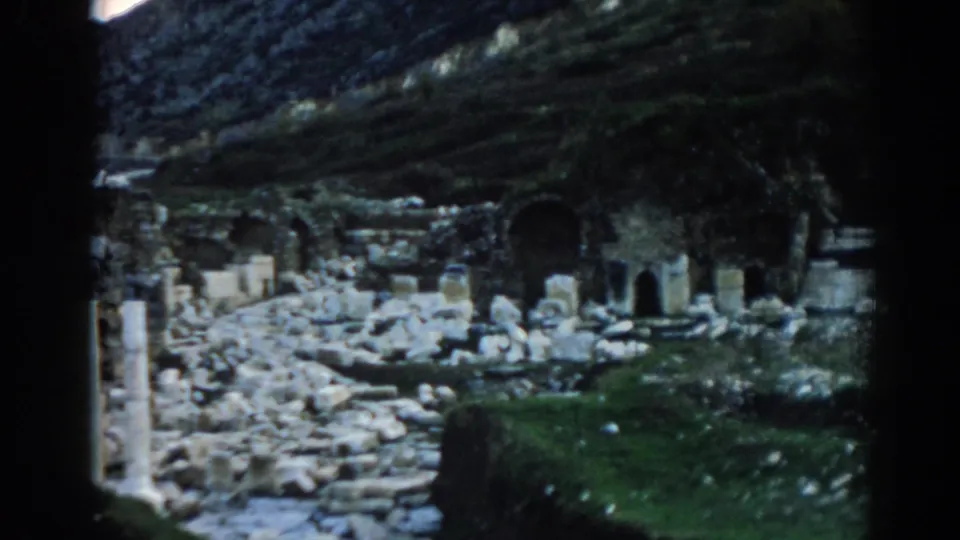 1962 visitor-shot frame at Ephesus showing scattered ruins, upright columns, dark archways, grass, and a sloping rocky backdrop.