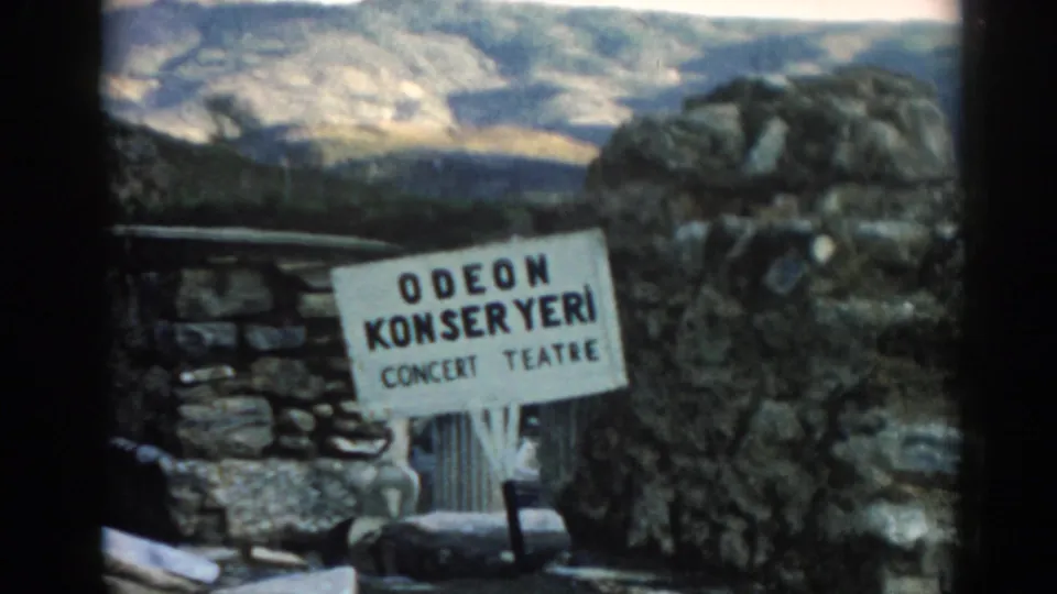 Visitor-shot 8mm frame at Ephesus showing the Odeon Konser Yeri sign against stone ruins and a pale hillside.