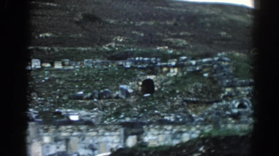 Ephesus home-movie frame showing open ruins, scattered stones, a dark opening, and hillside terrain beyond.