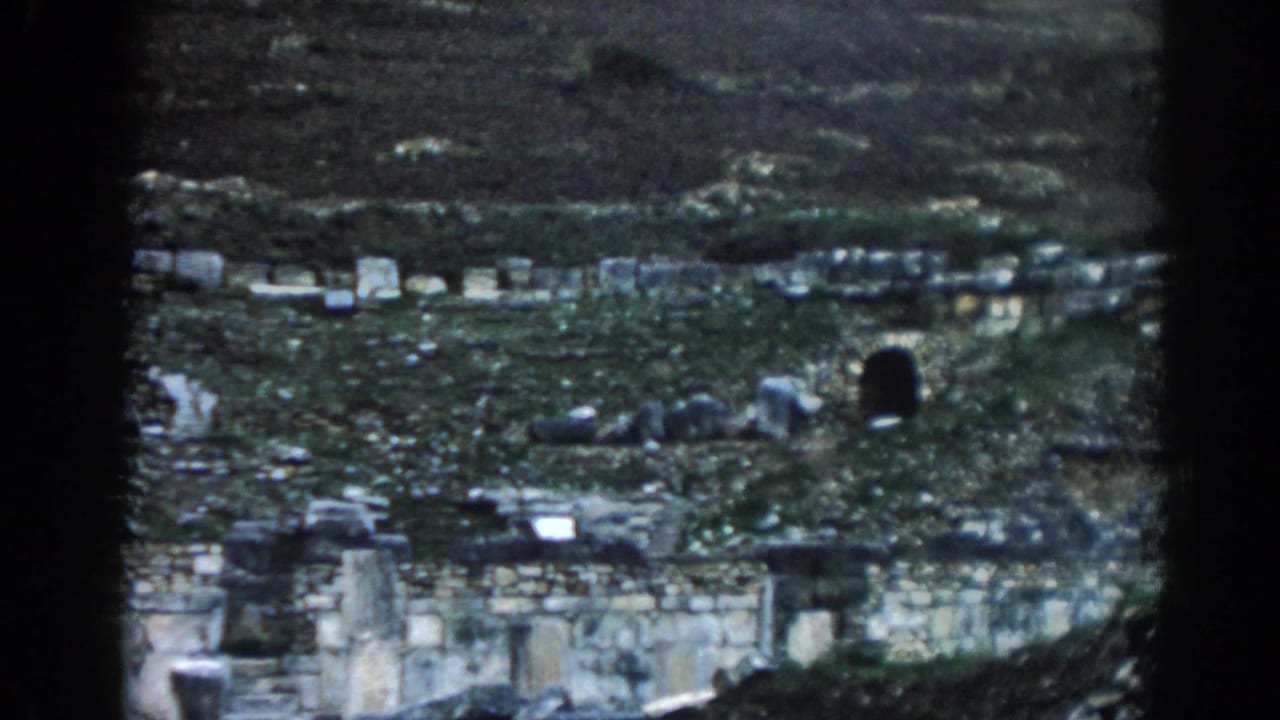 Visitor-shot 8mm frame from Ephesus, Turkey, with low ruins, uneven masonry, green vegetation, and a dark arched recess in the hillside.
