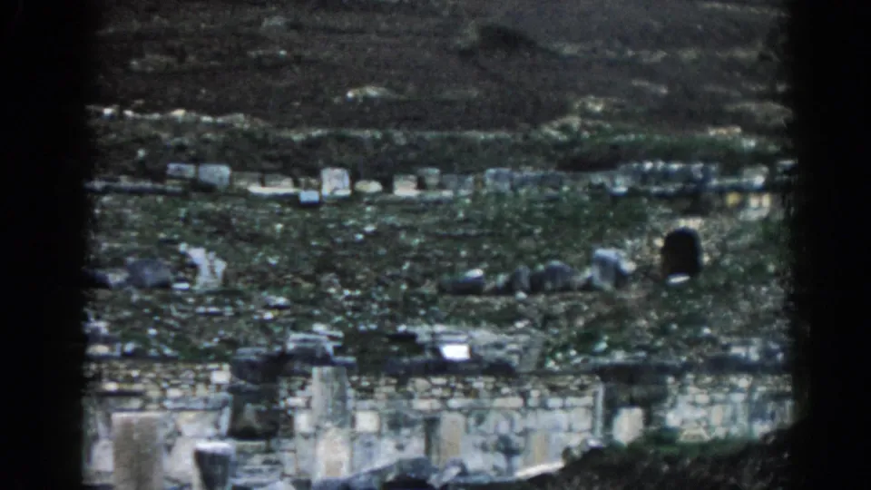 1962 home-movie frame of Ephesus ruins, with overgrown walls, scattered masonry, a dark opening, and heavy 8mm edge vignetting.