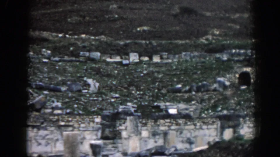 Ephesus, Turkey ruins in a 1962 8mm frame, showing worn stone walls, grassy ground, hillside texture, and a dark arched opening at right.