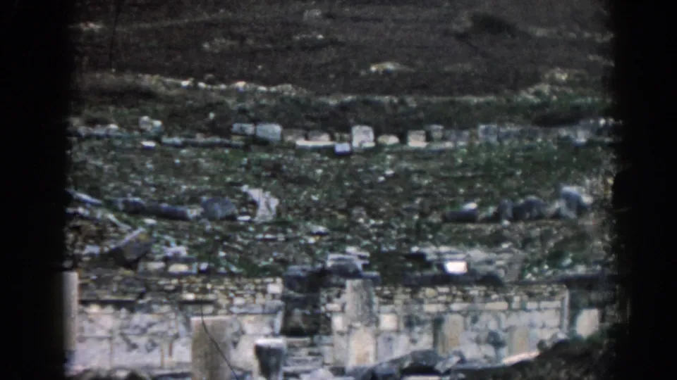 1962 Ephesus home-movie frame showing pale masonry, scattered stones, and a grey-green terraced hillside under muted light.