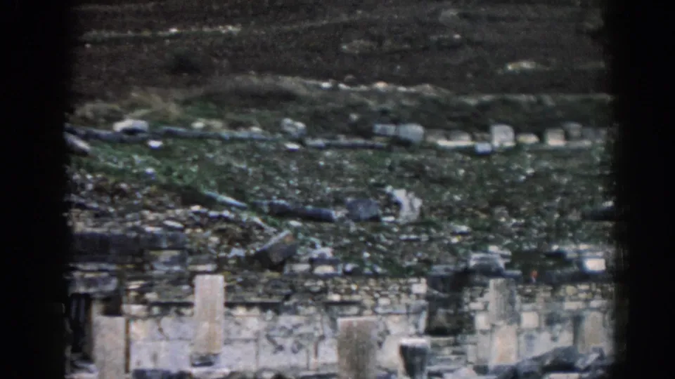 1962 Ephesus home-movie frame showing terraced ruins, pale wall sections, dark openings, and scattered hillside stones.