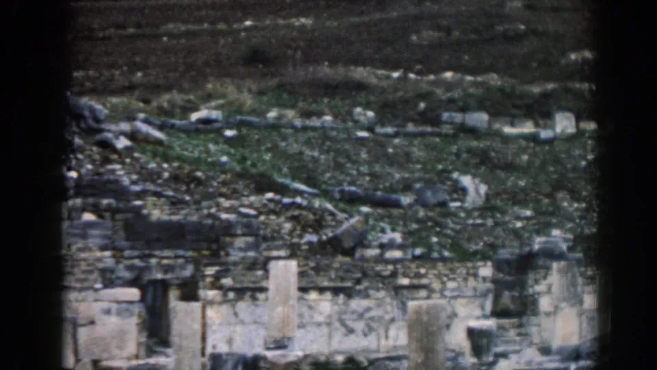 1962 Ephesus frame showing pale stone walls, broken masonry, and scattered rocks below a dark grey-green hillside.