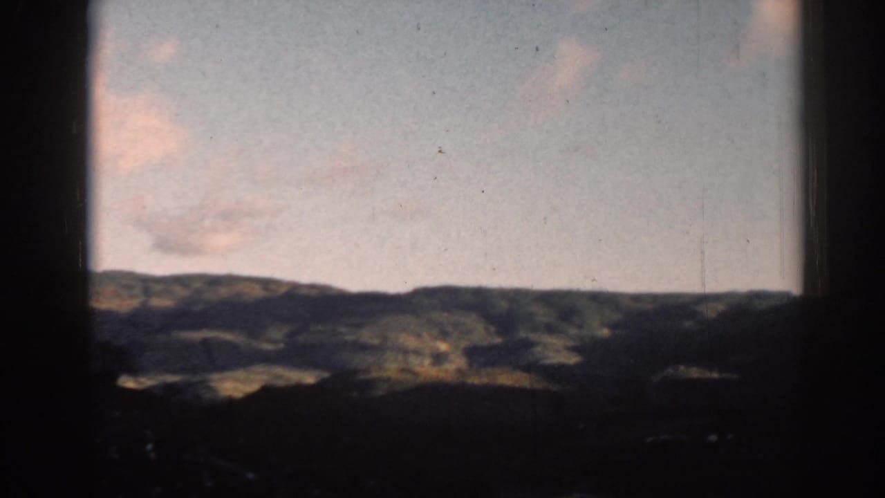 Dark window-framed view of rolling hills beneath a pale cloudy sky.