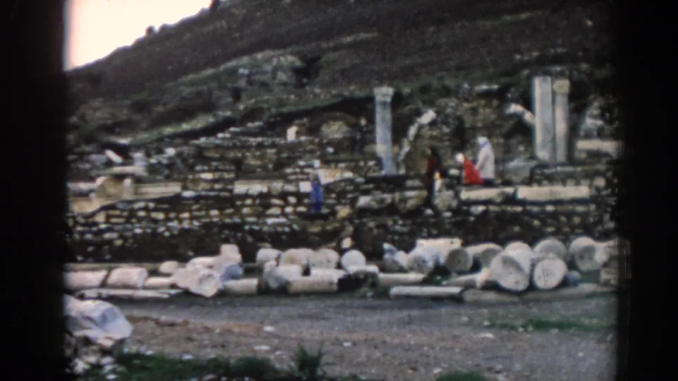 Wider 1962 Ephesus frame showing scattered stone fragments, standing columns, arched ruin elements, and visitors crossing the archaeological site.
