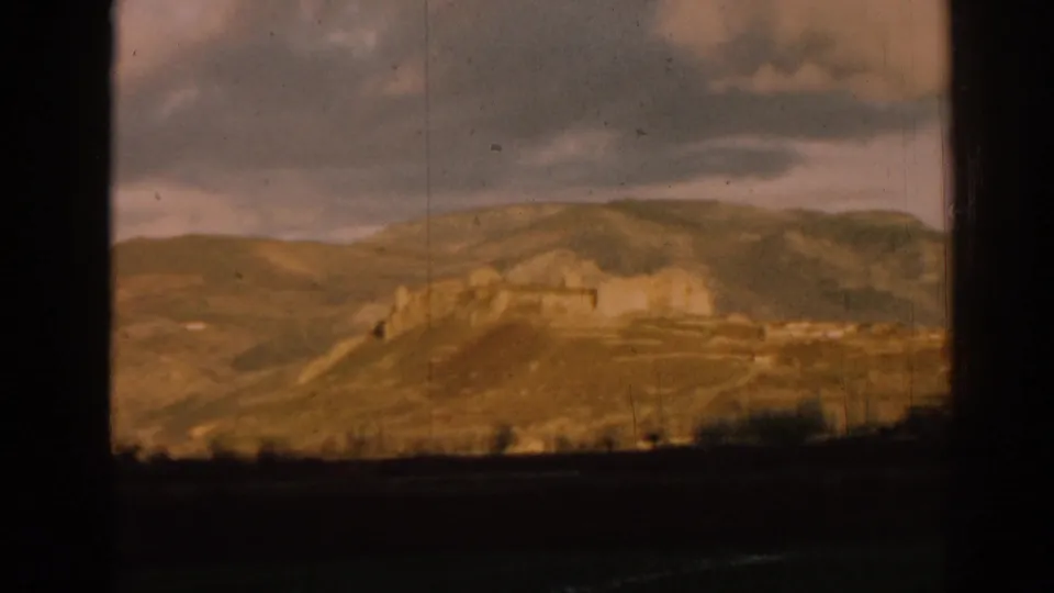 Wide 1962 Ephesus travel view of hillside ruins under a cloudy sky, with grain, scratches, and slight blur from 8mm film.