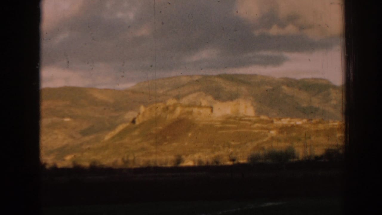 Distant ruins and hills seen through dark foreground framing under a cloudy sky.