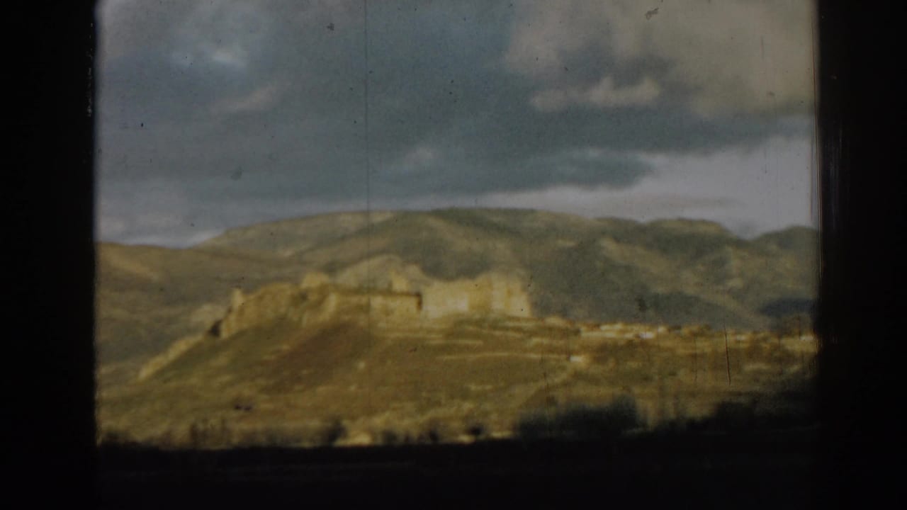Distant ruins on a hillside beneath a cloudy sky, framed by dark film borders.