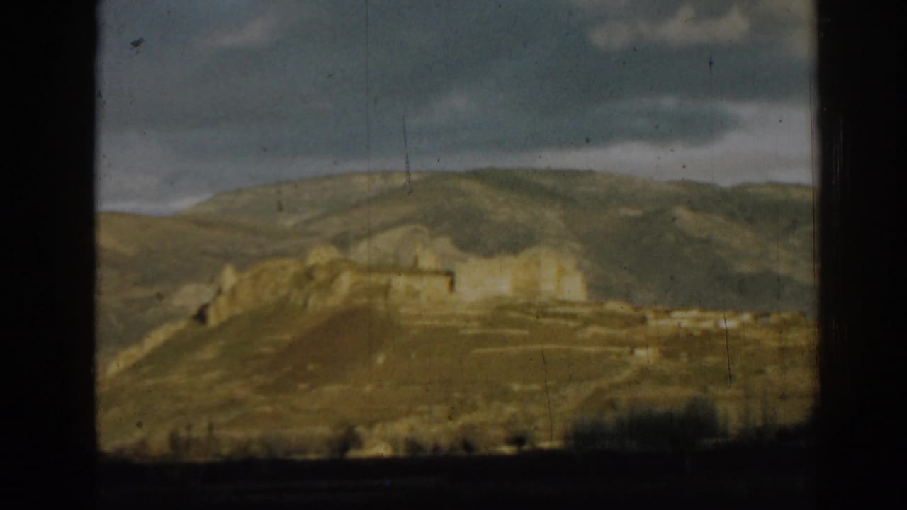Ruins stretch across a hillside with mountains and clouds behind them.
