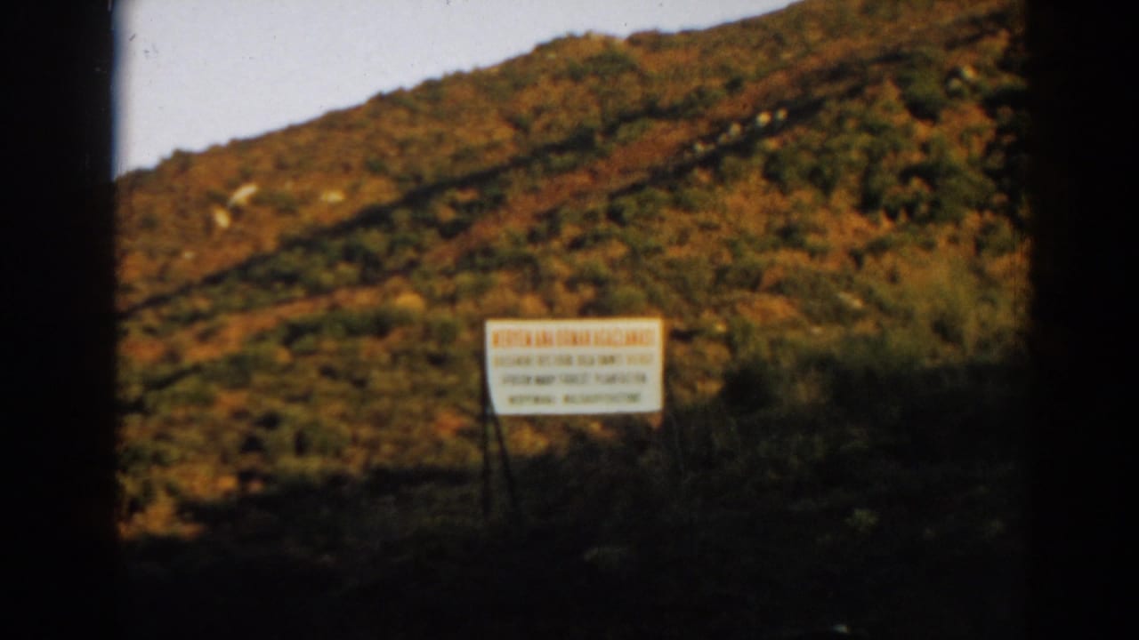 White sign with red lettering at the foot of a brown-green hillside.