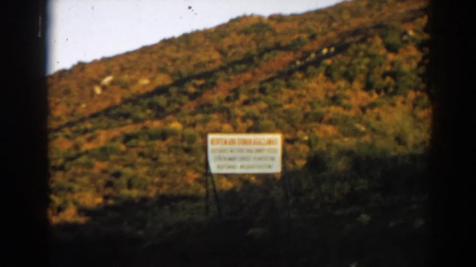 Final 1962 Ephesus-area 8mm frame with the white sign centered low against a warm brown and green hillside.
