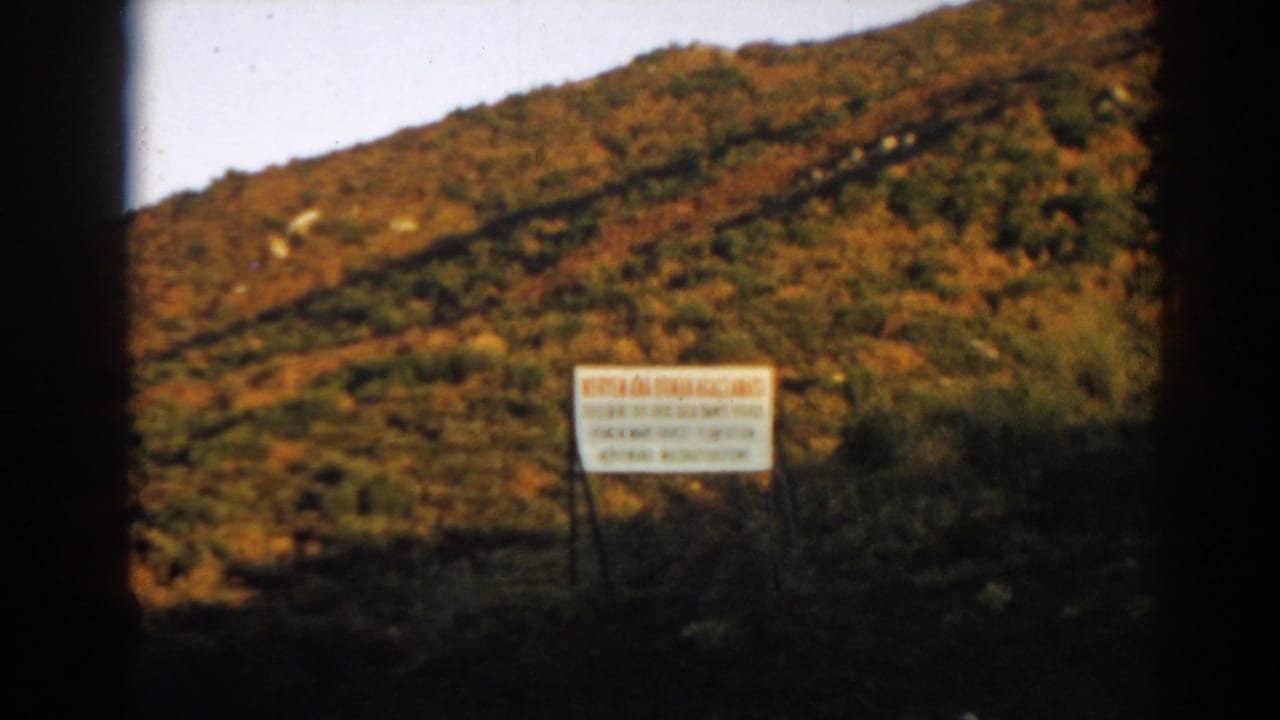 Sunlit green and brown hillside with a white sign seen through dark frame edges.