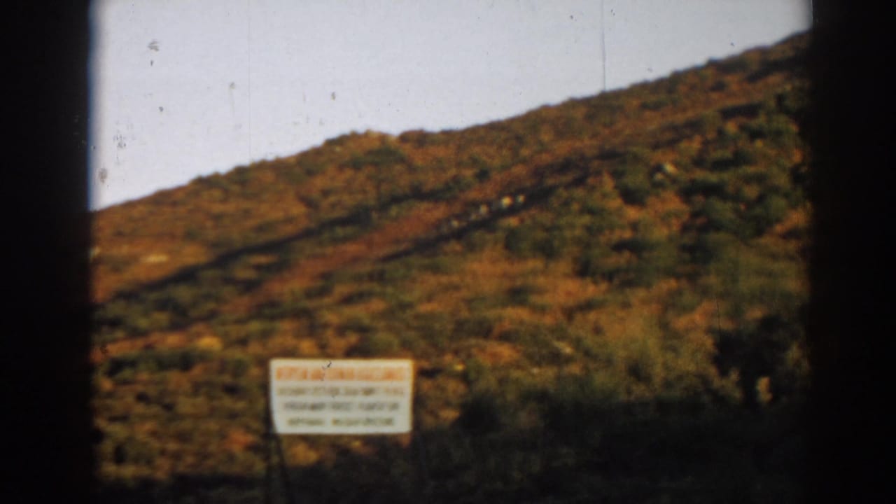 Visitor-shot 8mm view in Ephesus, Turkey, with a white sign below a sunlit scrub hillside and visible film dust.