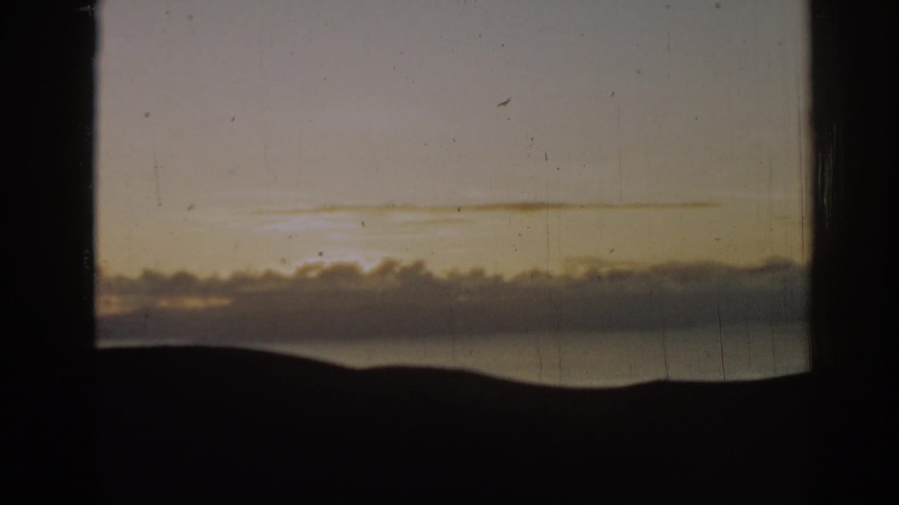 Window-framed sunset sky over a dark horizon and water with puffy clouds.