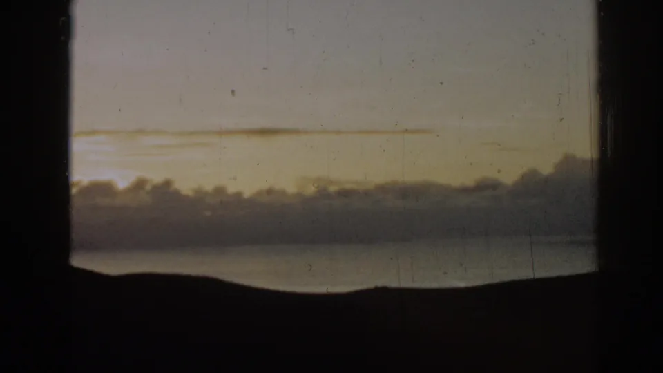 Vintage 8mm frame from the 1962 Ephesus, Turkey footage showing muted gold sky, puffy clouds, water, and a silhouetted horizon.