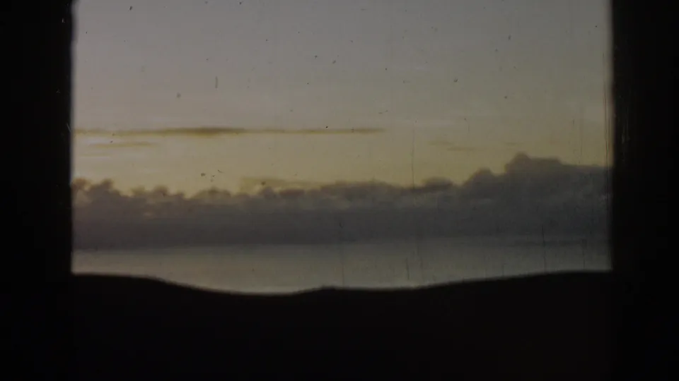 1962 8mm home-movie frame from Ephesus, Turkey, with a quiet dusk sky, puffy cloud bank, water, and silhouetted landform.