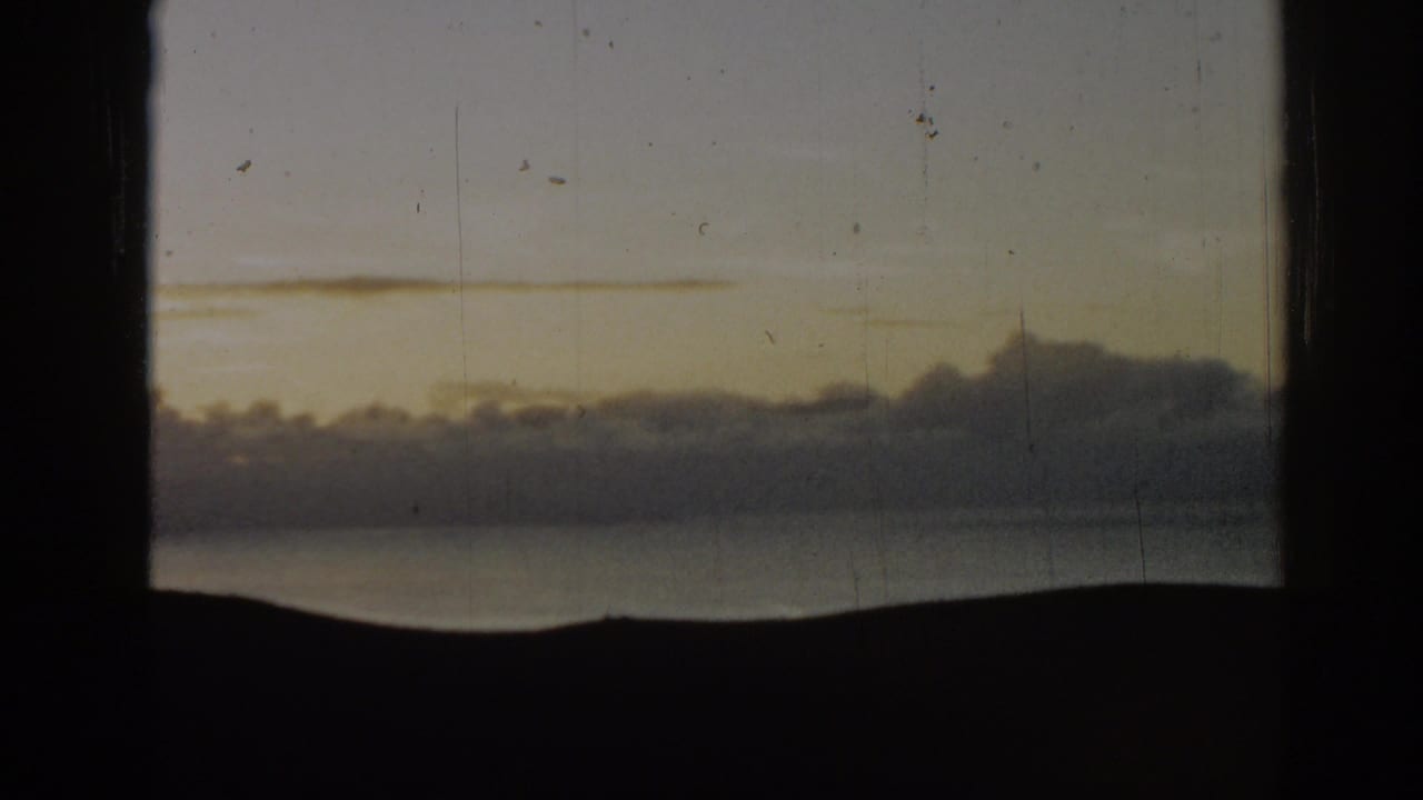 Pale sky and low clouds above a dark mountain silhouette and calm water, framed by black 8mm film borders.