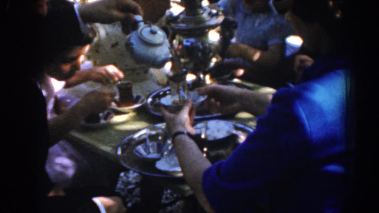 Hands gather around tea glasses and metal trays on a patterned tablecloth.