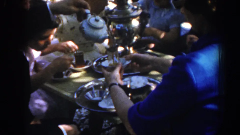 1962 Ephesus, Turkey home-movie frame showing guests' hands near clear tea glasses, saucers, trays, and a samovar or teapot on a tabletop.