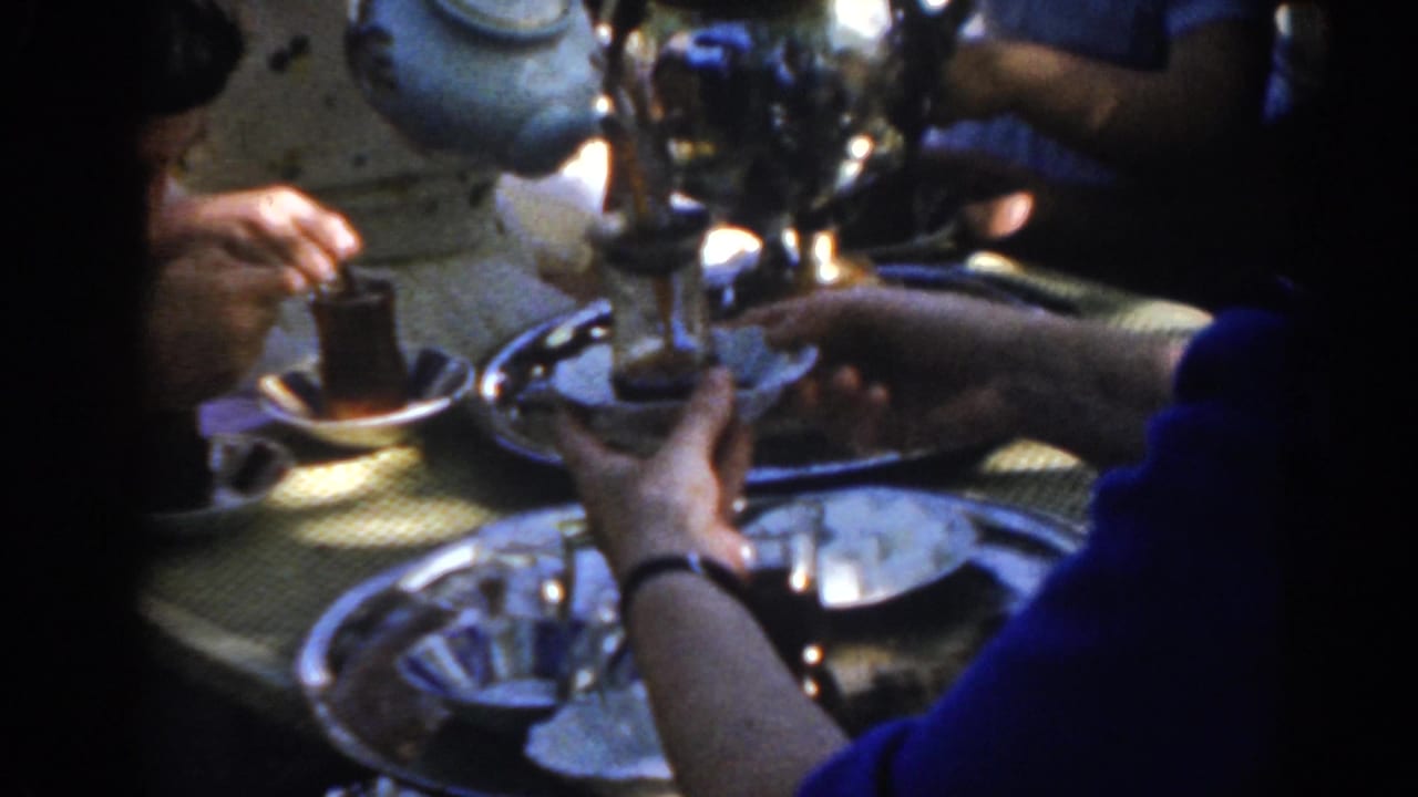 Tea glasses, trays, and a pale teapot or samovar are clustered on the table.