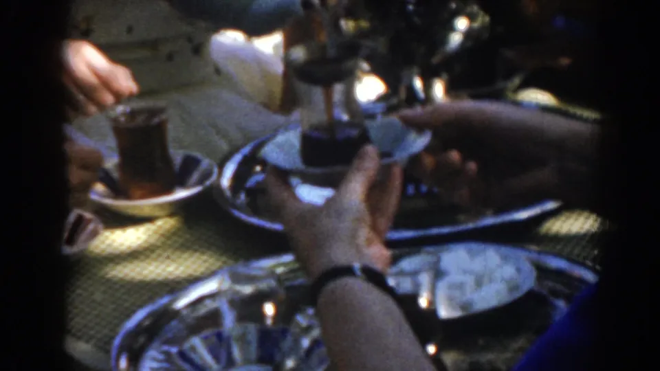 1962 Turkish home-movie close-up showing a hand holding a small clear tea glass near saucers, trays, and a metal serving vessel.