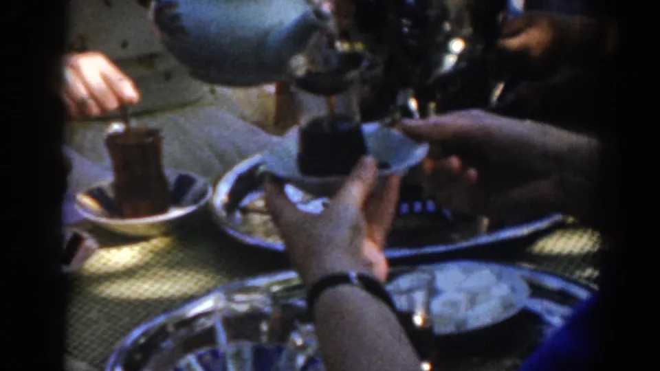 Ephesus, Turkey home-movie frame from 1962 showing dark tea in a small clear glass, hands, trays, and a pale samovar or teapot.
