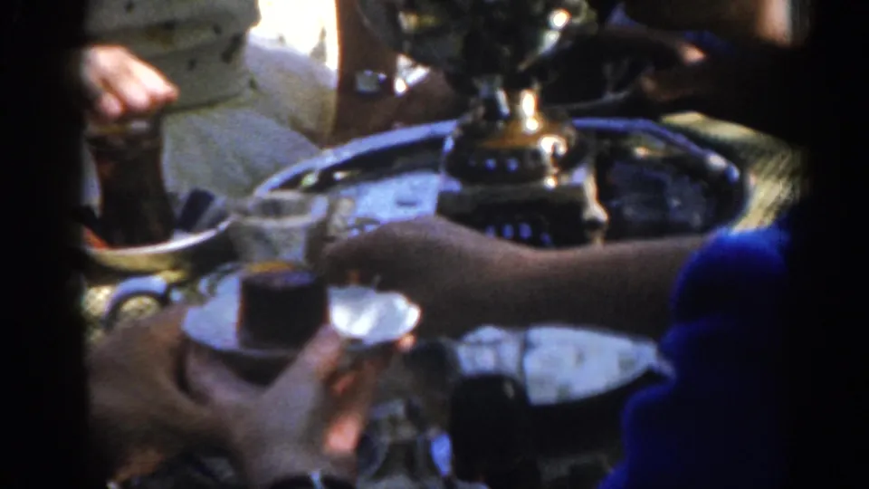 A blue-sleeved arm reaches across a tray of small glasses and a shiny pot during a 1962 gathering in Ephesus, Turkey.