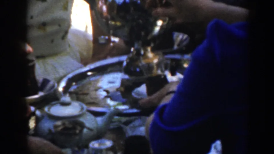 A person in a blue shirt leans in to serve drinks near a metal tray, small glasses, and a pot in 1962 Ephesus footage.