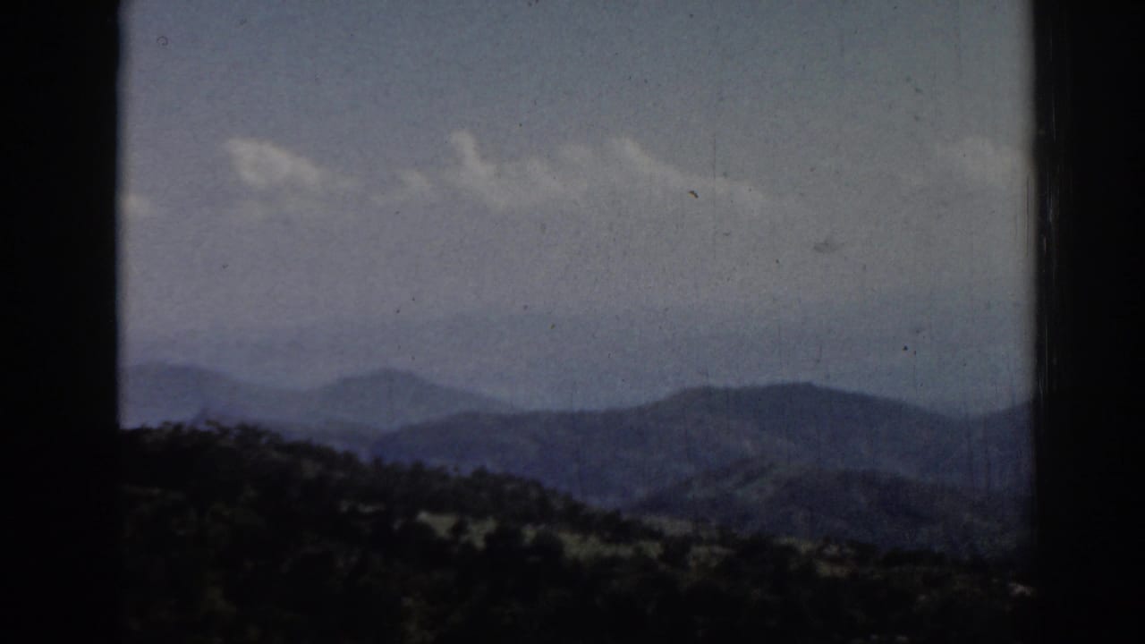 1962 Ephesus travel footage frame with rolling mountain forms, dark foreground vegetation, scattered clouds, and visible film dust.