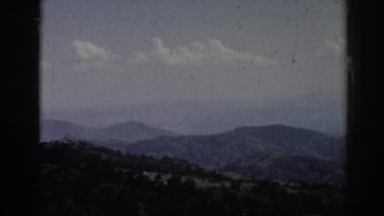 1962 8mm frame associated with Ephesus, Turkey, showing blue-gray mountain layers, a wooded lower ridge, clouds, and film wear.