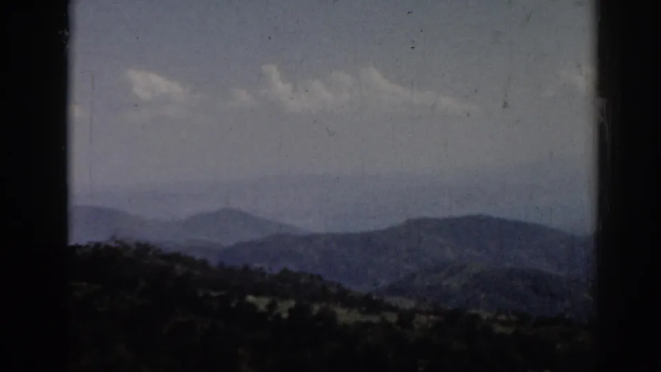1962 8mm frame associated with Ephesus, Turkey, showing blue-gray mountain layers, a wooded lower ridge, clouds, and film wear.